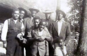A black-and-white photograph of a multigenerational family standing together outdoors in a grassy park beneath tall trees. One man near the centre wears a white hat. The group poses closely together for the camera.