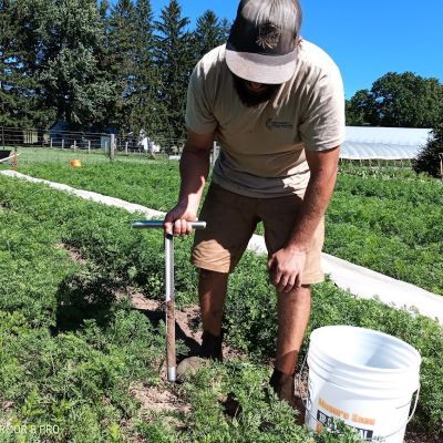 Jesse Way stands in foreground of photo in sand coloured shorts and a light t shirt, wearing a ball cap and looking down at the soil nitrate analysis sample he is collecting from a field of vegetables