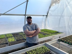 Arnest Sebbumba, a smiling Black farmer, stands in his greenhouse in front of some trays of seedlings