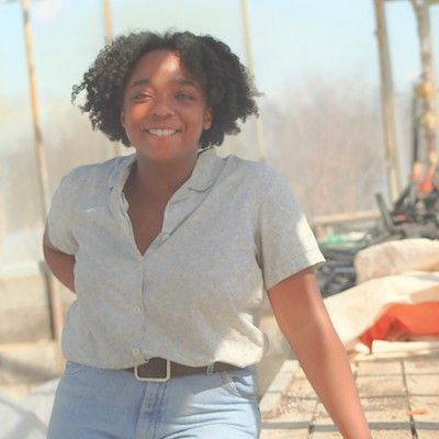 Farmer Aaliyah Fraser stands smiling at the camera inside a greenhouse on a sunny morning