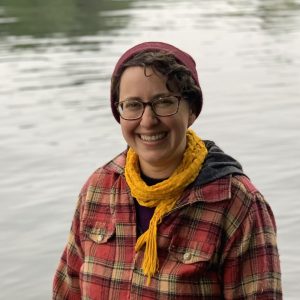 A headshot of Rebecca Ivanoff, with a big smile, wearing a red plaid shirt and beanie, in front of a body of water
