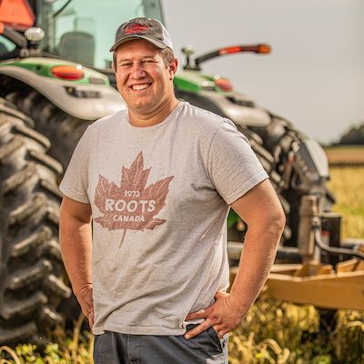 Brett Israel stands smiling with hands on hips in front of his green tractor, with a golden field in the background
