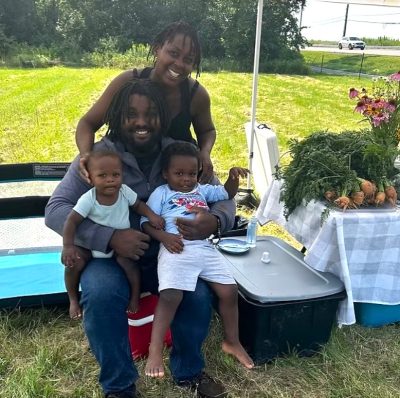 A smiling Black family of farmers under a market tent on a sunny day