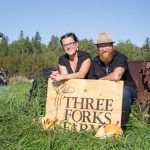 Two happy farmers crouch in a green field leaning on their wooden sign that reads "Three Forks Farms"