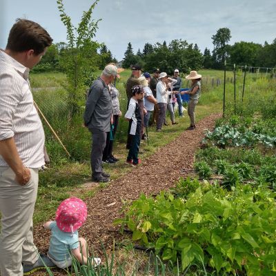 a group of people stand in a pathway facing a field of various crops. A child in a pink hat sits in the bottom left corner of photo, face turned toward a farmer wearing a wide brimmed hat.