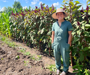 Shiying Lu, a smiling Chinese farmer in coveralls and a straw hat, stands in front of some amaranth on a sunny day