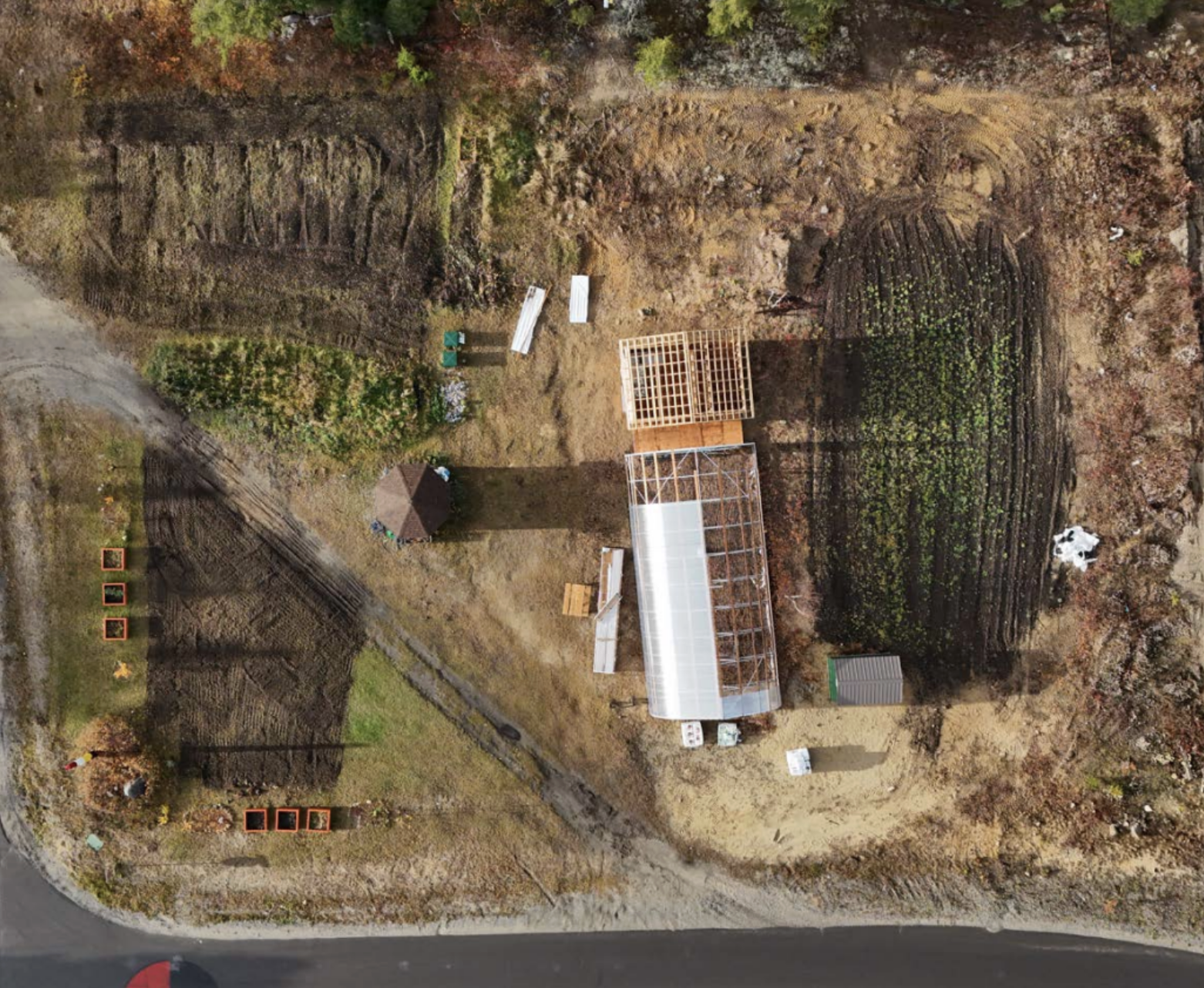 Aerial image of Leading Clouds Garden, on the right side of the plot are clearly defined rows of planted crops. Towards the center, there is a greenhouse. Adjacent to it is an open wooden frame. On the left side, a mix of bare earth and smaller green patches are visible, with some raised beds or garden boxes arranged neatly in a cluster. At the top left, are various garden tools or supplies.