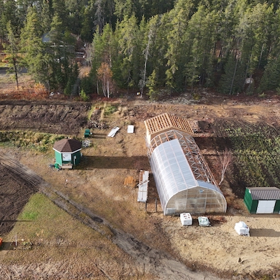 Aerial image of Leading Clouds Garden, on the right side of the plot are clearly defined rows of planted crops. Towards the center, there is a greenhouse. Adjacent to it is an open wooden frame. On the left side, a mix of bare earth and smaller green patches are visible, with some raised beds or garden boxes arranged neatly in a cluster. At the top left, are various garden tools or supplies.