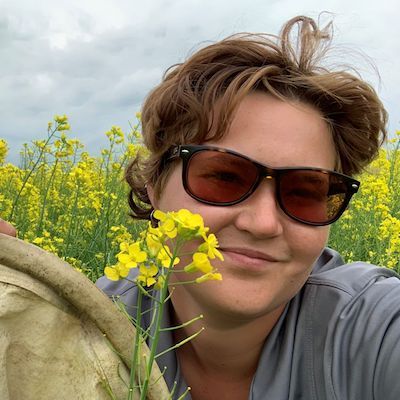 Lyss Gingrich smiles at the camera while crouching in a field of yellow-blooming mustard