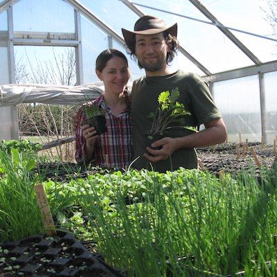 Farmers Alex & Kim of Mulberry Moon farm stand smiling in their hoop house in front of trays of green seedlings