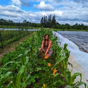 Rav crouches mid-frame, with a sundress and long hair. She is smiling in a row of corn and squash with a blue sky and white clouds behind her.