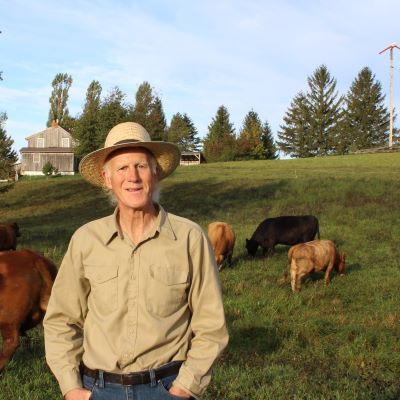 Tony stands in the foreground, wearing dark jeans, a beige button up, and a wide brimmed hat. He is smiling. Behind him, there are cattle grazing on grassland and a treeline of pines.