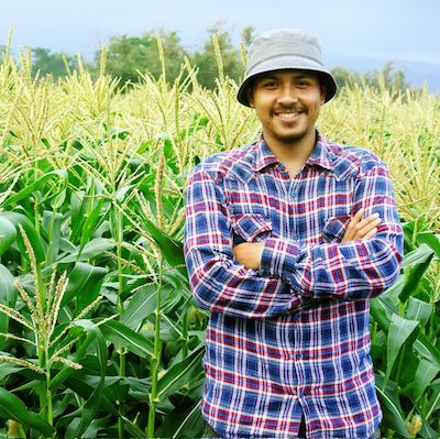 A person with light brown skin, wearing a grey brimmed hat and a blue and red plaid shirt stands in front of a lush green corn field, smiling