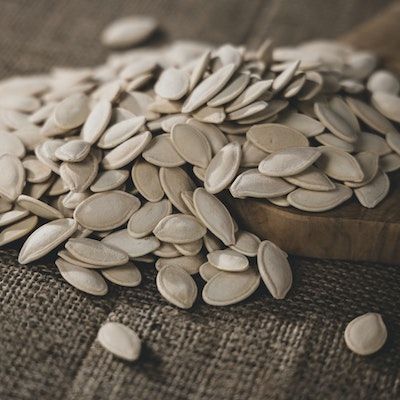 A pile of white pumpkin seeds spill onto a table covered by a brown burlap cloth