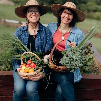 Stephanie is seated on the left, and Heather on the right. They are both wearing denim pants, and wide brimmed hats. The background is a green field. Stephanie has a basket of colourful produce in her lap including tomatoes, and Heather has a basket featuring garlic and kale. Both are smiling and Heather has an arm wrapped around Stephanie.