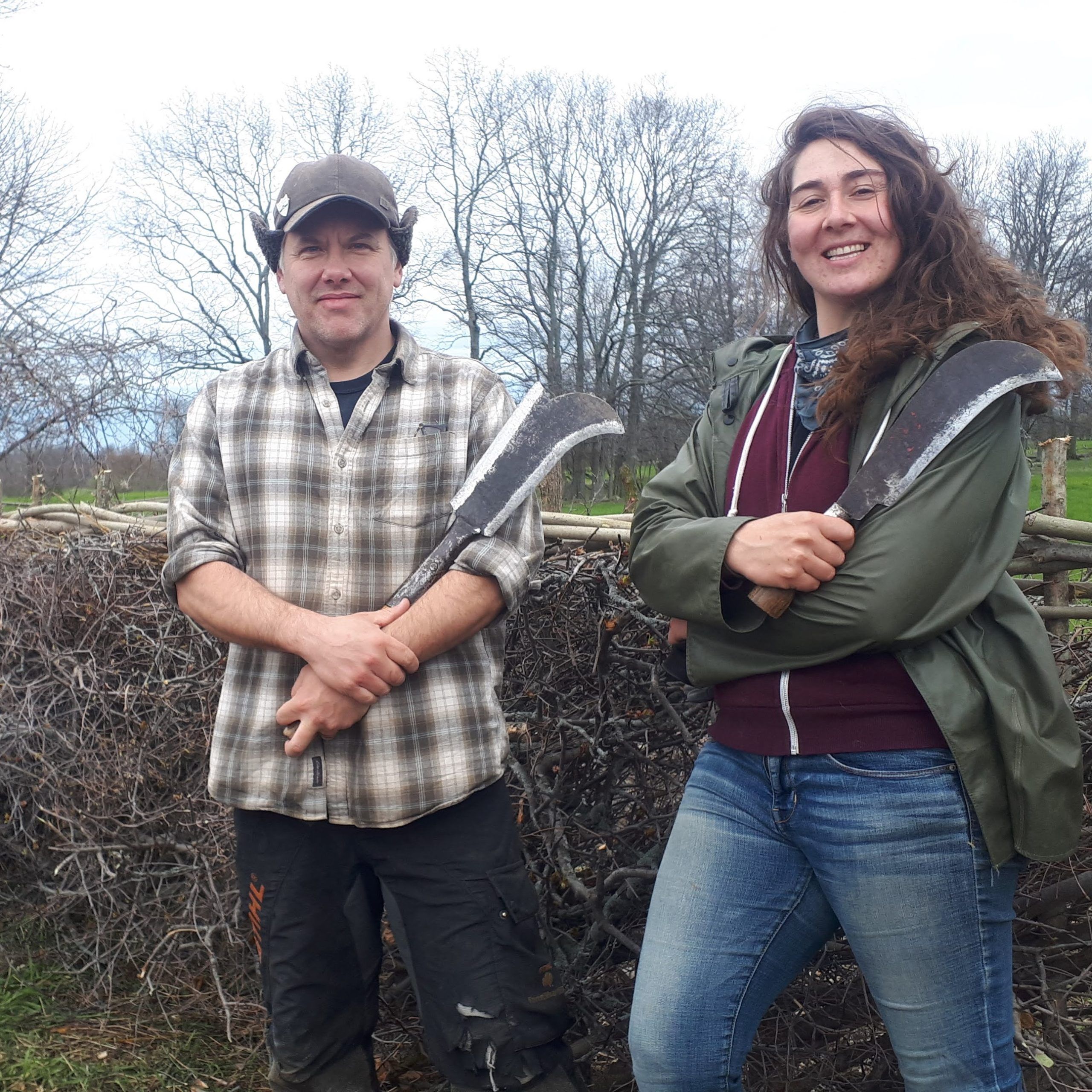 Two people stand posed with machetes smiling with a hedgerow in the background
