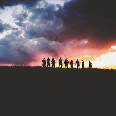 The silhouettes of 9 people stand atop a hill with a colourful sunset sky behind them